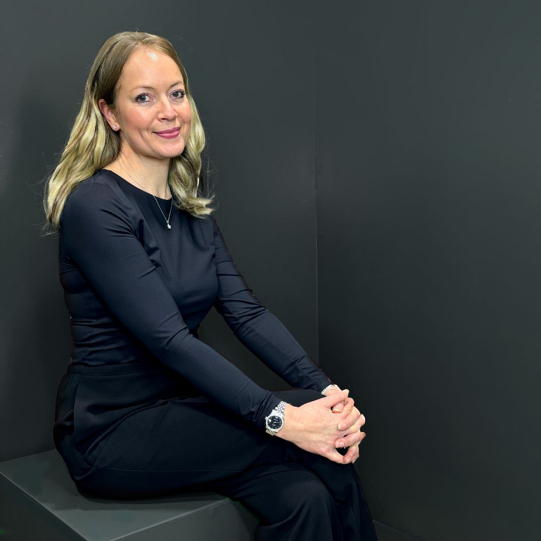 Lucy Pittaway seated against a dark background, wearing a long-sleeved black top and watch, smiling towards the camera.